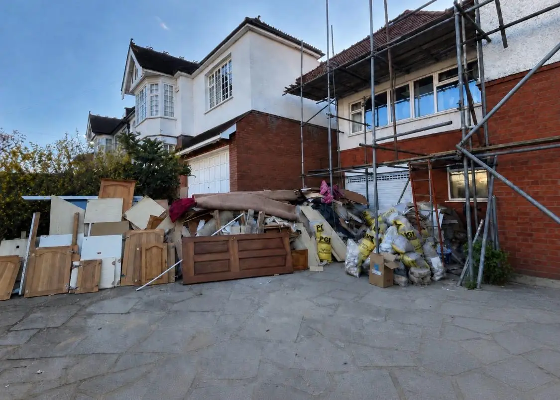 A house driveway part way through a clearance, internal doors and sheets of ply stacked on edge, rolled carpet and filled rubble bags across the flagstones, scaffolding on the house behind
