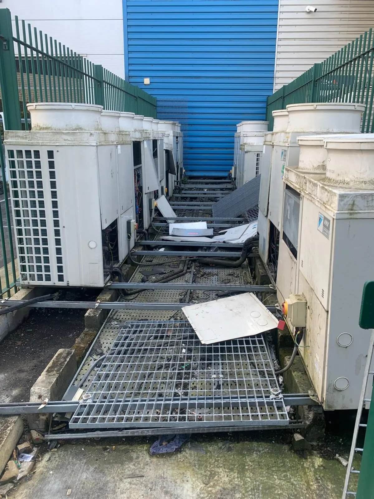 Before. A bank of air-conditioning condensers and old steel walkway grating behind the palisade.