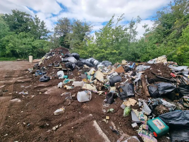 A large fly-tip of soil, black bags, plastic drums, cardboard and compost bags dumped on open ground, with trees and hedges behind