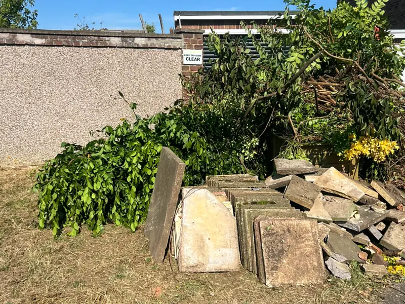 Hedge cuttings and cut branches heaped against a rendered garden wall, with a stack of old paving slabs leaning in front of them on dry grass