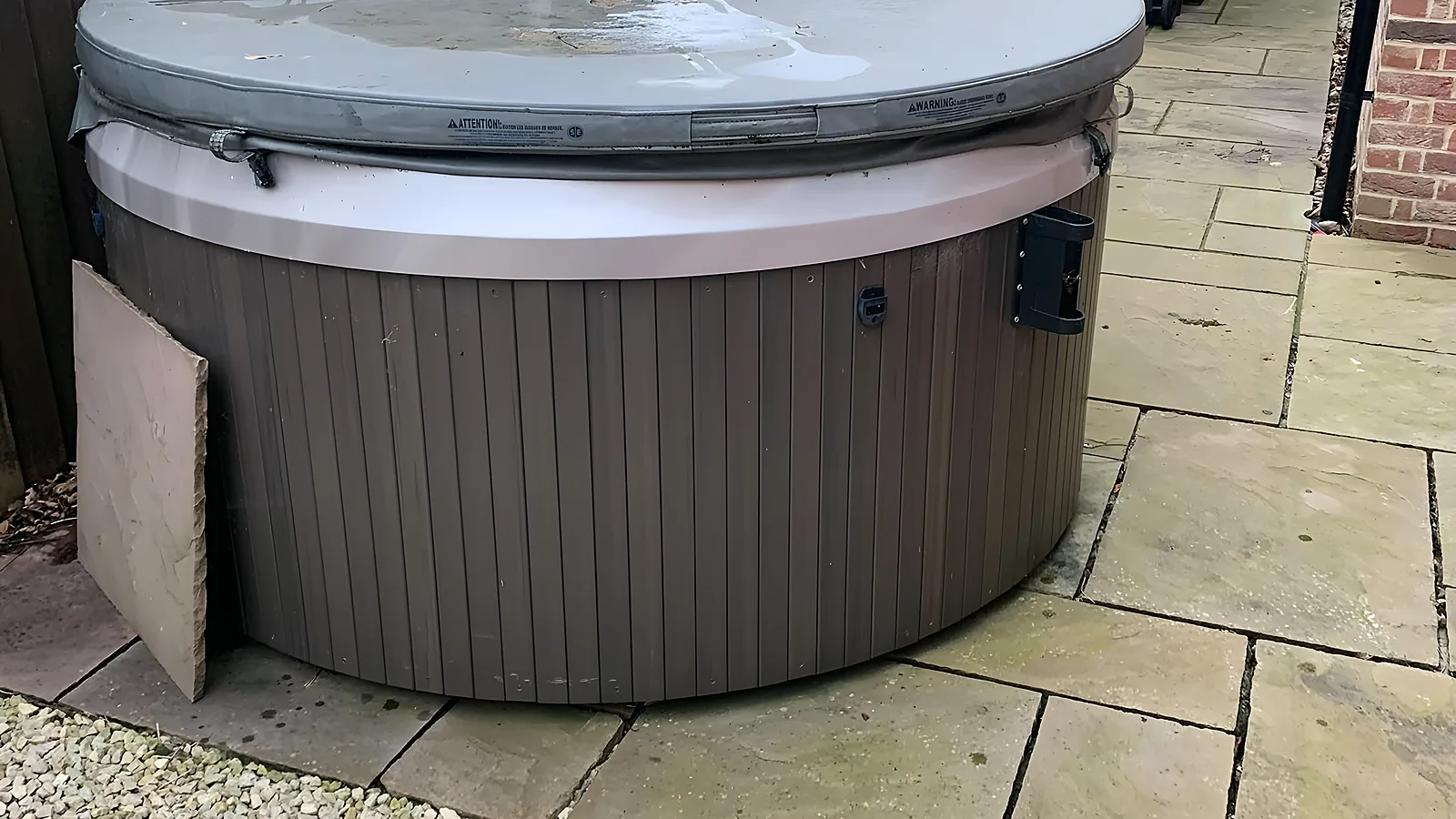 An old grey hot tub with its lid on, on a paved patio beside the wheelie bins